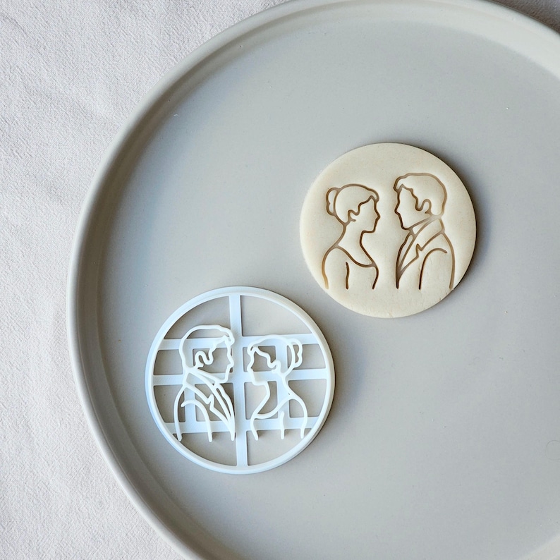 May include: A white plastic cookie cutter and a baked cookie with a couple design. The cookie cutter is round with a grid pattern. The baked cookie is round with a couple's silhouette.