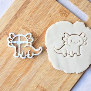 Puede incluir: Un cortador de galletas de axolotl de plástico blanco junto a un corte de galleta del mismo diseño. El axolotl tiene una cara sonriente y branquias rizadas. El cortador de galletas está sobre una tabla de cortar de madera.