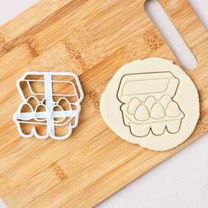May include: A white plastic egg carton cookie cutter and a cookie cut into the shape of an egg carton with six eggs. The cutter and cookie are on a wooden chopping board.