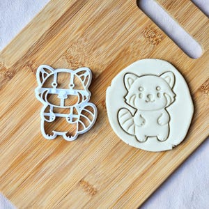 May include: A white plastic cookie cutter in the shape of a cartoon red panda, next to a cookie cut-out of the same design. The cutter is on a wooden cutting board.