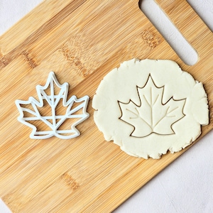 May include: A white plastic maple leaf cookie cutter next to a maple leaf imprint in dough. The cookie cutter is on a wooden cutting board. The dough is light beige in color.