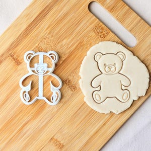 May include: A white plastic teddy bear cookie cutter and a piece of dough with a teddy bear imprint on a wooden board. The cookie cutter is next to the dough. The dough has a teddy bear shape pressed into it.