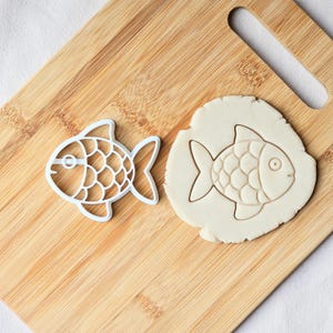 Puede incluir: Un cortador de galletas blanco en forma de pez junto a una galleta con forma de pez cortada en masa. El cortador tiene un patrón de escamas detallado. La galleta de masa tiene un contorno simple del pez.