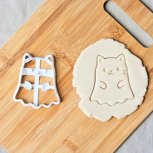 May include: A white plastic cat-shaped cookie cutter and a cookie cut-out on a wooden board. The cutter has a cat face and body shape. The cut-out shows the cat's face and body. The background is a light-coloured surface.
