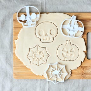 May include: A wooden chopping board with Halloween cookie cutters and dough. The white plastic cutters include a skull, a pumpkin, a spider web, and a spider. The dough shows the impressions of the cutters.