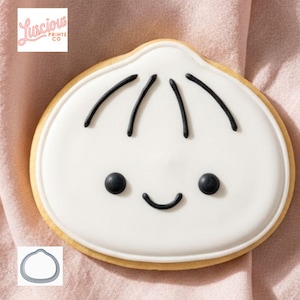 May include: A round sugar biscuit decorated with white icing and black icing details forming a simple face with a small smile and hair lines. The biscuit features a rounded top shape and is accompanied by a small metal biscuit cutter of the same shape.