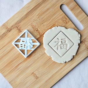 Könnte beinhalten: Ein weißer Kunststoff-Ausstecher in Diamantform mit chinesischen Schriftzeichen, neben einem Keks mit dem gleichen Design. Der Ausstecher und der Keks liegen auf einem Holzschneidebrett.