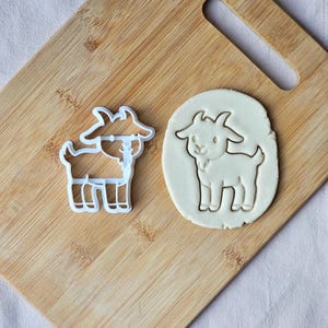 May include: A white plastic goat-shaped cookie cutter and a goat-shaped cookie on a wooden board. The cutter has a detailed goat outline. The cookie is light brown with a goat shape pressed into it. The board is made of wood.