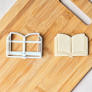 May include: Two book-shaped items on a wooden surface. One is a white plastic cookie cutter, and the other is a beige cookie. Both are open book designs. The cookie cutter is a hollow outline, while the cookie has detailed pages.