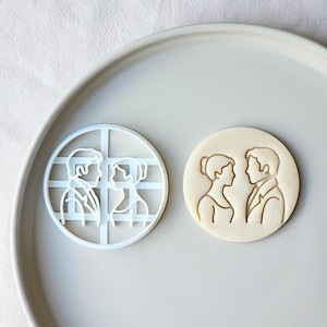 May include: A white plastic cookie cutter and a baked cookie with an embossed design of a couple facing each other. The cookie cutter is round with a grid pattern. The baked cookie is light brown.