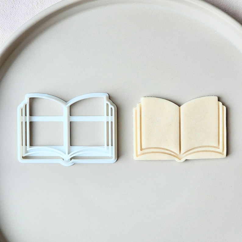 May include: A light blue plastic cookie cutter in the shape of an open book sits next to a baked cookie in the same shape. The cookie is light beige with brown lines to represent the pages.