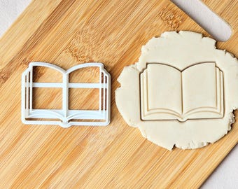 May include: A white plastic cookie cutter shaped like an open book with a central spine and two pages, positioned next to a piece of light-colored dough that has been pressed with the same book-shaped design to create a clear, indented impression.