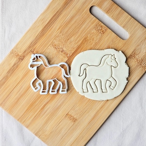 Op de afbeelding: Een witte, paardvormige koekjessnijder naast een paardvormige uitsparing in deeg. De snijder staat op een houten snijplank. Het paard wordt afgebeeld in een wandelhouding, met gedetailleerde benen en manen.