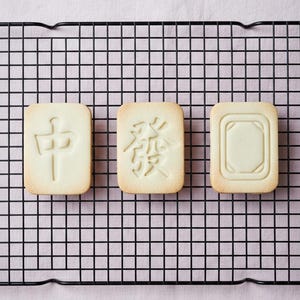 May include: Three rectangular cookies with embossed designs, arranged on a black wire rack. The cookies have different Chinese characters and a geometric pattern. The background is a light pink fabric.