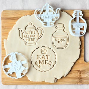 May include: A collection of white plastic cookie cutters in various shapes, including teapots, bottles, and a circular design with the words "EAT ME." The cutters are pressed into a sheet of dough, creating impressions of the designs. The text "WE'RE ALL MAD HERE" and "DRINK ME" are also visible.