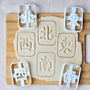 May include: A wooden cutting board with cookie dough and white plastic cookie cutters. The cutters are in the shape of Chinese characters, with impressions in the dough. The characters are 北, 東, 西, and 南.