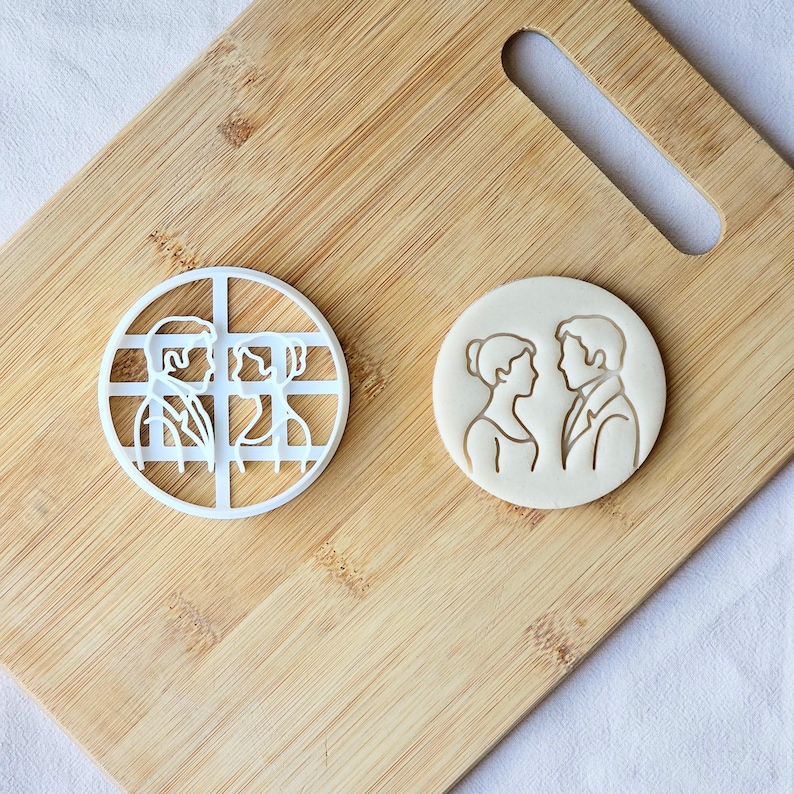 May include: Two cookie cutters on a wooden cutting board. One is a white plastic circular cutter with a grid design and a couple. The other is a round cookie with a couple's silhouette.
