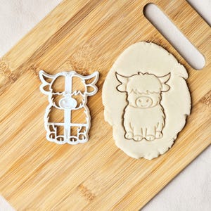Può includere: Un tagliabiscotti in plastica bianca a forma di mucca delle Highlands accanto a un biscotto a forma di mucca delle Highlands su un tagliere di legno. Il biscotto è marrone chiaro.