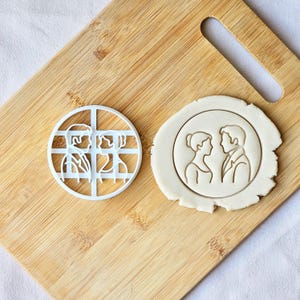 May include: A white plastic cookie cutter with a circular design of a couple facing each other. Next to it is a round cookie with the same design pressed into it. The cutter is on a wooden cutting board.