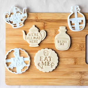 Peut inclure: Un ensemble d'emporte-pièces et de biscuits sur le thème d'"Alice au pays des merveilles". Les emporte-pièces sont en plastique blanc en forme de théière, de bouteille et de forme circulaire. Les biscuits sont beiges et portent les phrases "We're All Mad Here", "Drink Me" et "Eat Me".