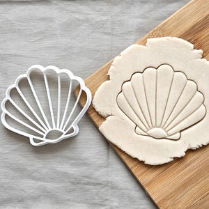 Op de afbeelding: Een witte schelpvormige koekjesuitsteker naast een stuk deeg met de schelpvorm erin gedrukt. De uitsteker is gemaakt van wit plastic. Het deeg ligt op een houten snijplank. Bakken thema.