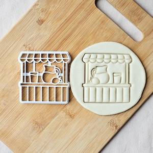 Könnte beinhalten: Ein weißer Ausstecher in Form eines Limonadenstandes mit Krug, Zitrone und Glas. Daneben ein Keks mit dem gleichen eingeprägten Design. Die Artikel liegen auf einem Holzschneidebrett.