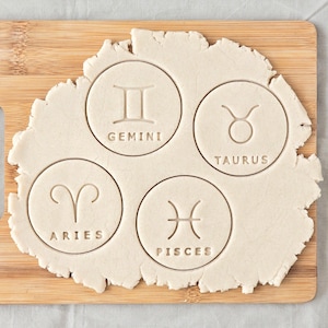 Pode incluir: Uma tábua de corte de madeira contém uma massa com quatro impressões circulares de signos do zodíaco: Gêmeos, Touro, Áries e Peixes. Os signos são gravados na massa, com os nomes de cada signo impressos abaixo.