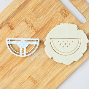 May include: A white plastic watermelon-shaped cookie cutter and a piece of dough with a watermelon slice imprint. The cutter is on a wooden chopping board. The dough has a watermelon slice imprint with seed details.