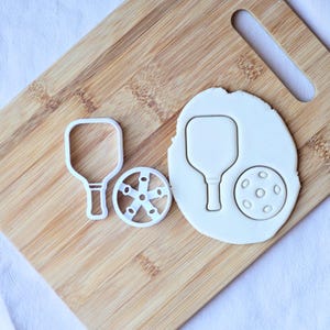 Könnte beinhalten: Weißer Pickleball-Ausstecher-Satz aus Kunststoff auf einem Holzbrett. Das Set enthält einen Schläger, einen Ball und einen radförmigen Ausstecher. Die Ausstecher werden zum Backen und Basteln verwendet.