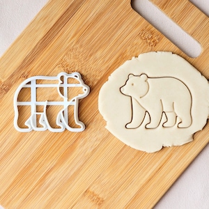 Puede incluir: Un cortador de galletas blanco con forma de oso junto a una huella de oso en masa. El cortador tiene un diseño de rejilla dentro del contorno del oso. La huella de la masa muestra la forma del oso. Ambos están sobre una tabla de cortar de madera.