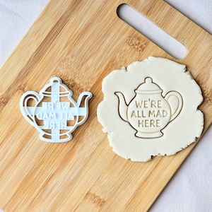 以下が含まれることがあります： 白いプラスチック製のティーポット型のクッキーカッターと、「WE'RE ALL MAD HERE」と刻印されたクッキー。 クッキーカッターは木製のまな板の上にあります。