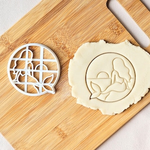 Puede incluir: Cortador de galletas de plástico blanco con forma de sirena junto a una galleta con la impresión de una sirena. La sirena está en un círculo con un fondo de puesta de sol. En una tabla de cortar de madera.