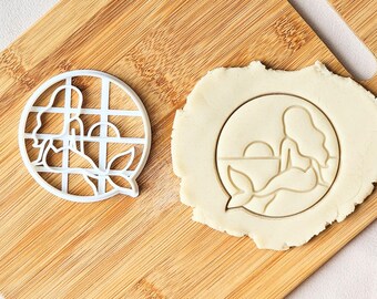 May include: A white circular cookie cutter featuring a mermaid silhouette sitting by a sunset, alongside a piece of rolled dough with the same mermaid and sunset design pressed into the surface, creating a clear outline of the scene.