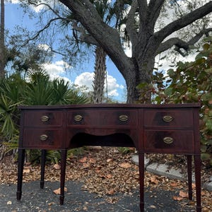 May include: A dark mahogany desk with six drawers and brass hardware. The desk has a curved apron and slender legs. The wood grain is visible, and the desk appears to be vintage or antique. The desk is outdoors, with a tree and foliage in the background.