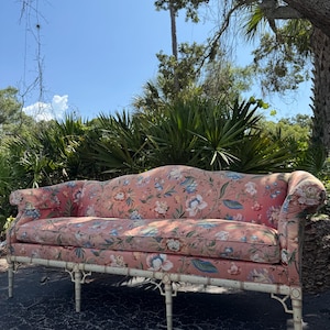 May include: A three-seat sofa featuring a salmon-pink fabric upholstery with a multi-colored floral pattern. The sofa has rolled arms, a curved backrest, and a light-colored wooden frame with a bamboo-style design and six vertical legs.