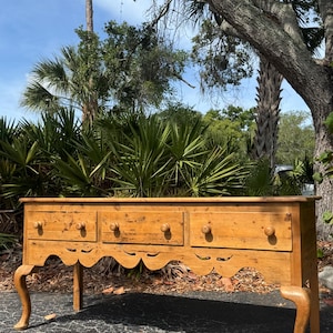 May include: A long, antique wooden dresser with three drawers and a scalloped apron. The dresser has turned legs and round wooden knobs. The wood has a warm, golden tone, and the piece appears to be outdoors.