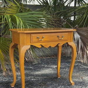 May include: A small, wooden side table with a single drawer and ornate brass hardware. The table has curved legs and a scalloped apron. The wood has a warm, golden-brown finish, and the table measures approximately 30 inches long.