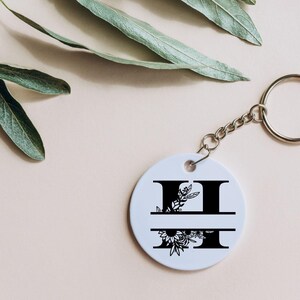 May include: White round keychain with a black floral design and the letter "H" split in half. The keychain has a silver metal ring.