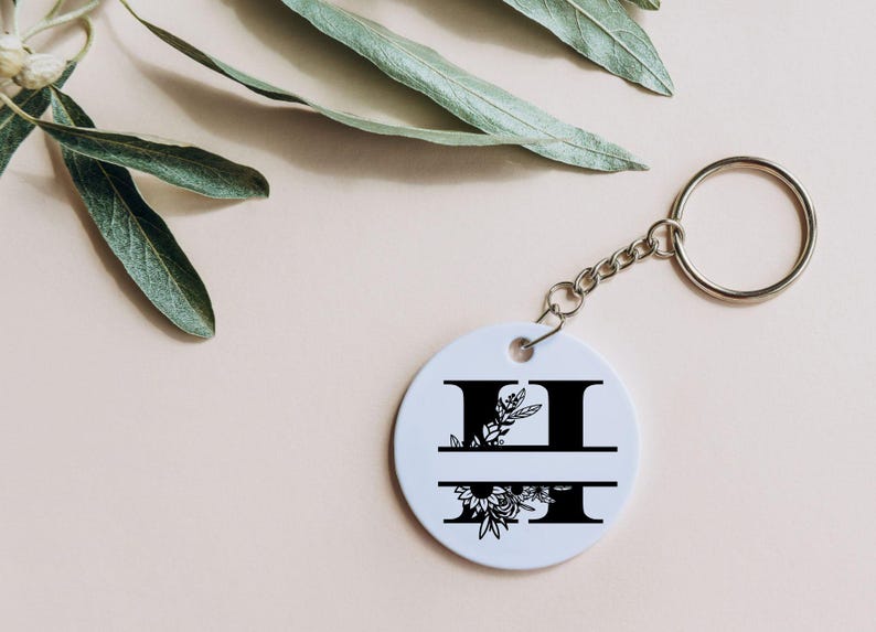 May include: White round keychain with a black floral design and the letter "H" split in half. The keychain has a silver metal ring.