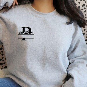 May include: A gray sweatshirt with a black floral design and the letter "D" on the front.