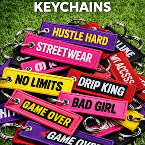 Könnte beinhalten: Eine Sammlung von individuell bestickten Schlüsselanhängern in verschiedenen Farben, darunter Pink, Lila, Gelb und Rot. Jeder Schlüsselanhänger zeigt auffällige Texte wie "Hustle Hard", "Streetwear" und "Game Over". Die Schlüsselanhänger liegen auf einer grünen, grasbewachsenen Oberfläche.