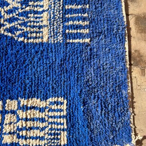 Op de afbeelding: Een blauw en wit gestreept tapijt met een geometrisch ontwerp. Het tapijt is gemaakt van een dik, getextureerd materiaal en heeft een franje rand.