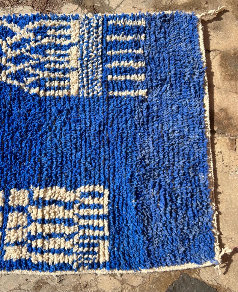 Op de afbeelding: Een blauw en wit gestreept tapijt met een geometrisch ontwerp. Het tapijt is gemaakt van een dik, getextureerd materiaal en heeft een franje rand.
