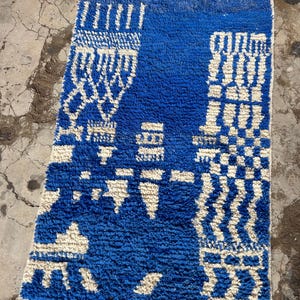 Op de afbeelding: Een blauw en wit Marokkaans tapijt met een geometrisch patroon. Het tapijt heeft een franje aan de onderkant.