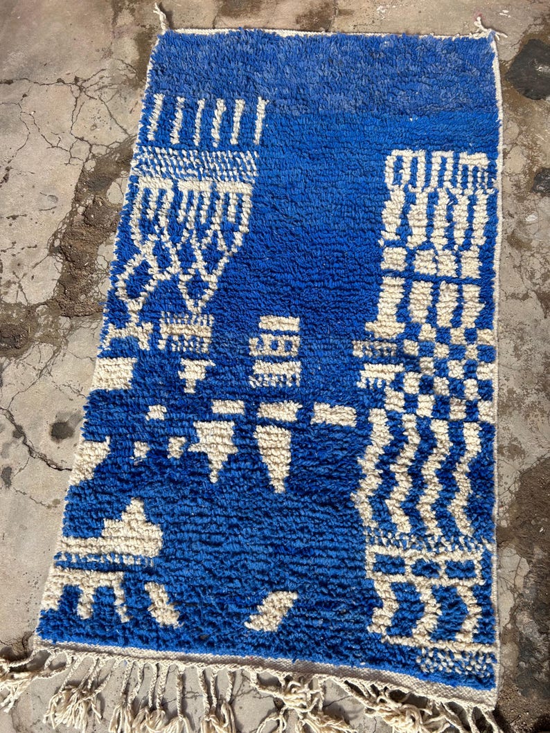 Op de afbeelding: Een blauw en wit Marokkaans tapijt met een geometrisch patroon. Het tapijt heeft een franje aan de onderkant.