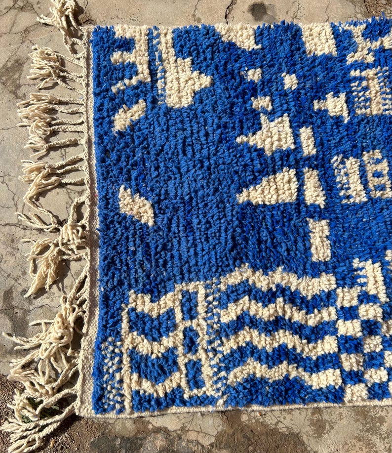 Op de afbeelding: Een blauw en wit geweven tapijt met een geometrisch patroon en franjes aan &eacute;&eacute;n kant.