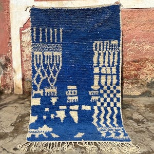 Op de afbeelding: Een blauw en wit Marokkaans tapijt met een geometrisch patroon. Het tapijt heeft een franje rand en is gemaakt van wol.