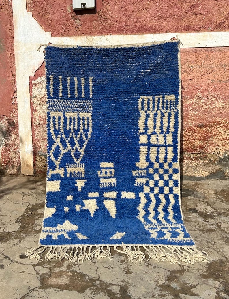 Op de afbeelding: Een blauw en wit Marokkaans tapijt met een geometrisch patroon. Het tapijt heeft een franje rand en is gemaakt van wol.