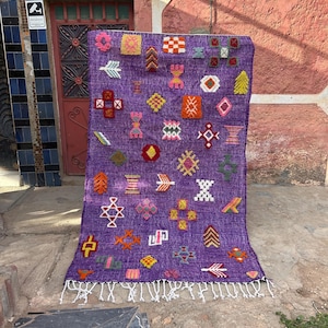 Op de afbeelding: Een paars tapijt met kleurrijke geometrische patronen en een witte franje. Het tapijt heeft verschillende vormen en ontwerpen in roze, geel, oranje en groen, waardoor een textiel ontstaat met een opvallende textuur.