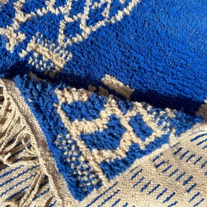 Op de afbeelding: Een blauw en wit Marokkaans tapijt met een geometrisch patroon en franjes. Het tapijt is gemaakt van wol en heeft een zachte, pluche textuur.